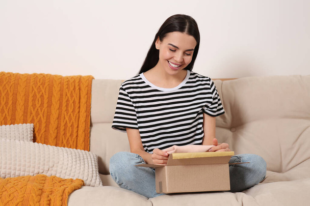 Happy young woman opening parcel on sofa at home. Internet shopping - Zdjęcie, obraz