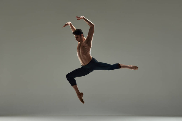 Young handsome man with muscular shirtless body, ballet dancer making performance over grey studio background. Concept of art, classical dance, inspiration, creativity, fashion, beauty, choreography - Photo, image
