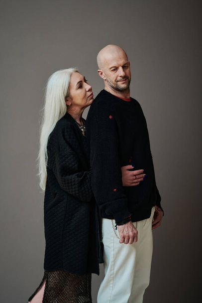 Vertical medium long studio portrait of loving mature woman with long gray hair hugging man from behind, gray background - 写真・画像