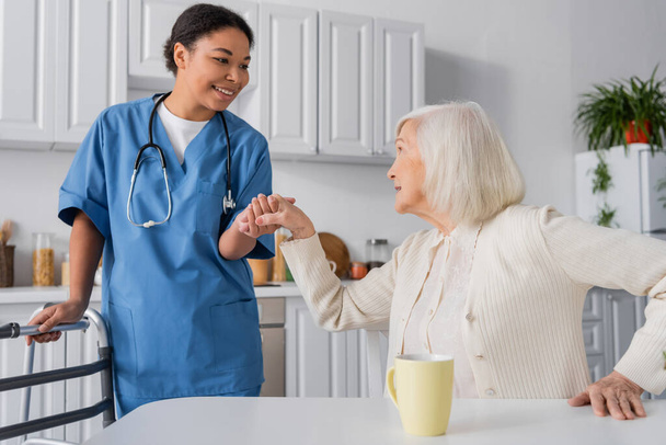 feliz enfermeira multirracial em azul uniforme segurando mão de mulher sênior com cabelos grisalhos enquanto estava perto walker  - Foto, Imagem