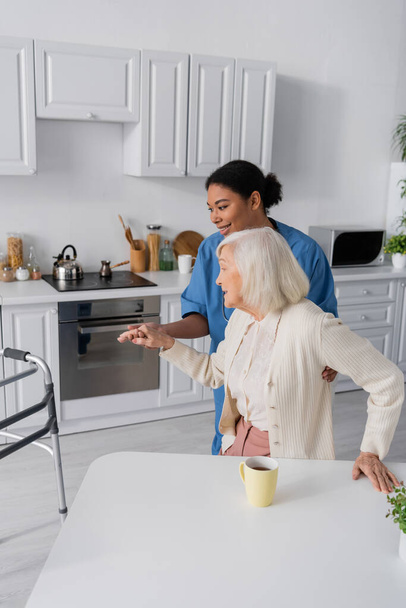 happy multiracial caretaker in blue uniform holding hand of senior woman with grey hair while standing near walker  - Фото, зображення