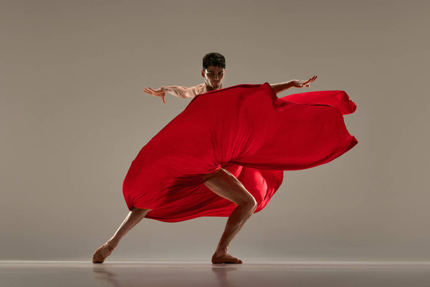 Modern ballet performance. Young, impressive, handsome man dancing with red silk fabric against grey studio background. Concept of art, classical dance, inspiration, creativity, fashion, choreography - Foto, Bild