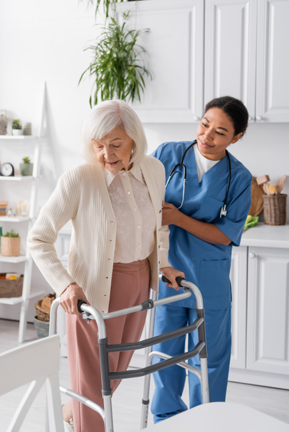 брюнетка багаторасова медсестра в синій формі, що підтримує старшу жінку з сірим волоссям ходити з допомогою ходунки  - Фото, зображення