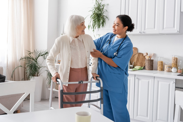enfermera multirracial feliz en uniforme azul que apoya a mujer mayor con el pelo gris que camina con la ayuda del caminante  - Foto, imagen