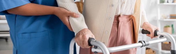 καλλιεργημένη άποψη της πολυφυλετικής νοσοκόμας με μπλε στολή που υποστηρίζει ηλικιωμένη γυναίκα περπατώντας με τη βοήθεια του περιπατητή, λάβαρο  - Φωτογραφία, εικόνα
