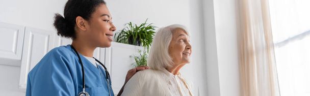 Mavi üniformalı neşeli, çok kültürlü hemşire evde gri saçlı mutlu yaşlı kadına bakıyor.  - Fotoğraf, Görsel