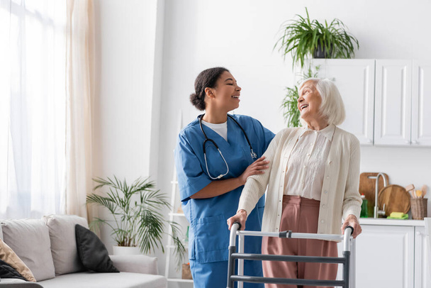 onnellinen monirotuinen sairaanhoitaja univormussa nauraa eläkkeellä olevan naisen kanssa seistessään lähellä kävelykehystä kotona  - Valokuva, kuva