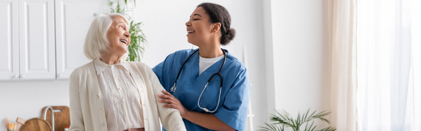 happy multiracial nurse in uniform laughing with retired woman at home, banner  - Фото, зображення