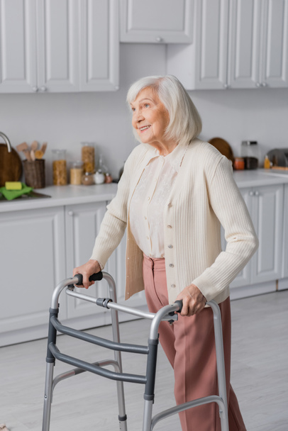 Positive Seniorin mit grauen Haaren geht mit Rollator zu Hause  - Foto, Bild