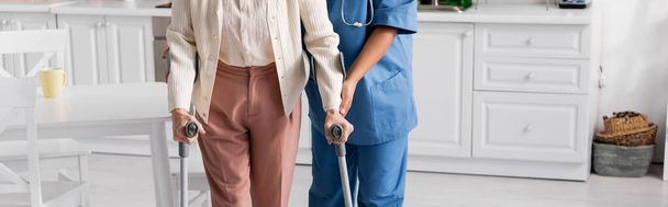 περικοπή άποψη της ηλικιωμένης γυναίκας χρησιμοποιώντας πατερίτσες, ενώ το περπάτημα κοντά νοσοκόμα στο σπίτι, πανό  - Φωτογραφία, εικόνα