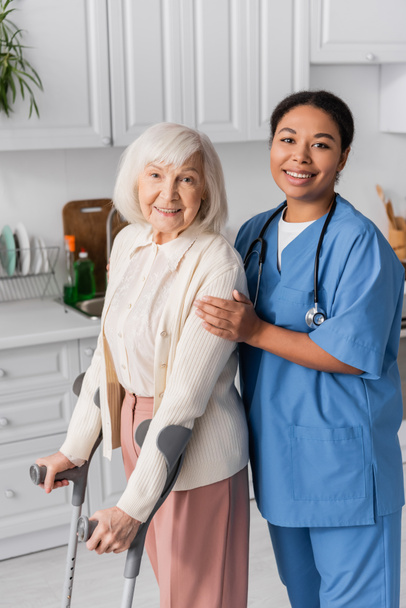 donna in pensione con i capelli grigi utilizzando stampelle e sorridente mentre si cammina vicino felice infermiera multirazziale a casa  - Foto, immagini