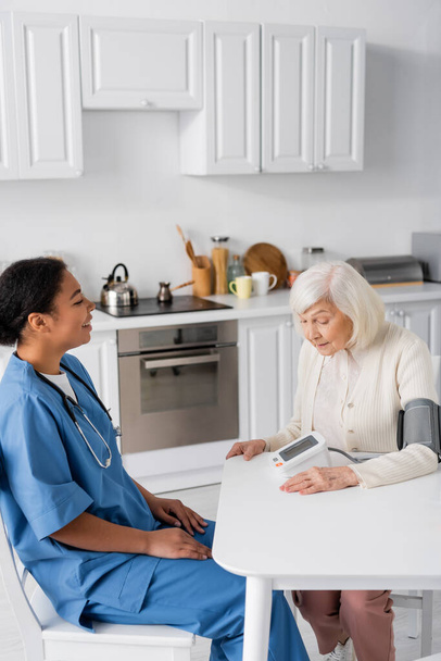 щаслива багаторасова медсестра дивиться на старшу жінку з сірим волоссям вимірюючи кров'яний тиск
  - Фото, зображення
