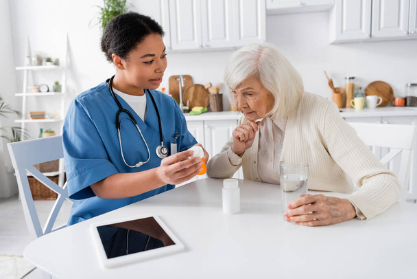 monirotuinen sairaanhoitaja osoittaa lääkitystä vanhempi nainen vieressä digitaalinen tabletti pöydällä  - Valokuva, kuva