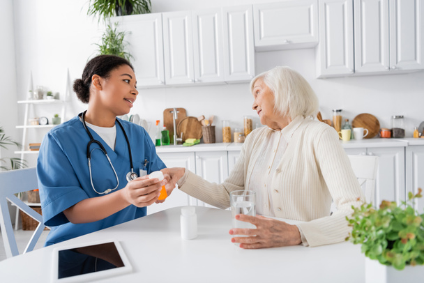multiracial nurse showing medication to skeptical senior woman next to digital tablet on table  - Foto, immagini