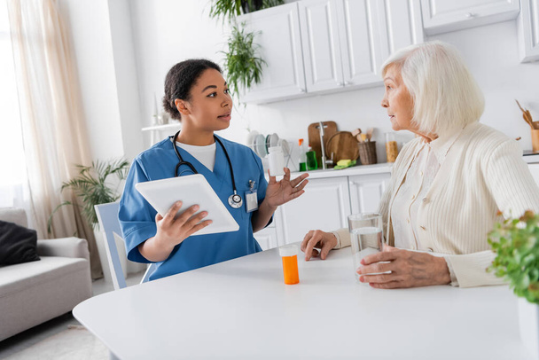 bruna infermiera multirazziale usando tablet digitale e tenendo i farmaci mentre parla con una donna anziana con i capelli grigi a casa  - Foto, immagini