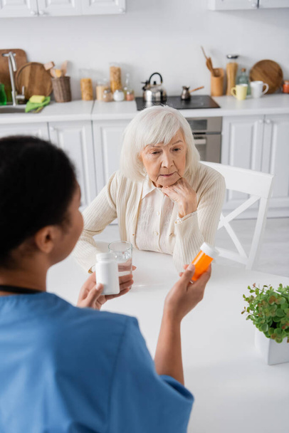 overhead weergave van brunette multiraciale verpleegkundige houden medicatie terwijl praten met gepensioneerde vrouw met grijs haar - Foto, afbeelding