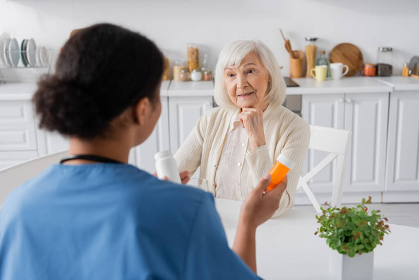 gepensioneerde vrouw met grijs haar op zoek naar multiraciale verpleegster met medicatie op wazig voorgrond  - Foto, afbeelding