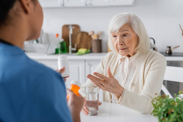 mujer jubilada con el pelo gris preguntando acerca de la medicación en manos de la enfermera multirracial en primer plano borroso  - Foto, Imagen