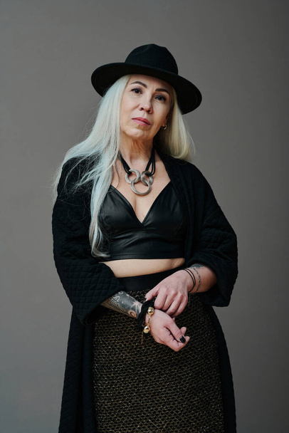 Vertical medium long studio portrait of attractive mature Caucasian woman with long gray hair wearing modern goth outfit looking at camera - Photo, Image