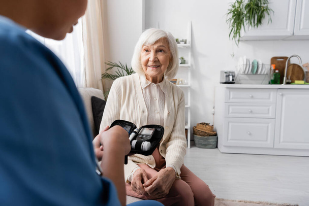 ευτυχισμένη ηλικιωμένη γυναίκα κάθεται στον καναπέ και ψάχνει σε πολυφυλετική νοσοκόμα κρατώντας κιτ διαβήτη  - Φωτογραφία, εικόνα