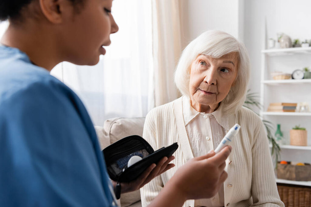 multiraciale verpleegkundige die lancet pen vasthoudt en uitlegt hoe deze te gebruiken aan oudere vrouw met grijs haar  - Foto, afbeelding