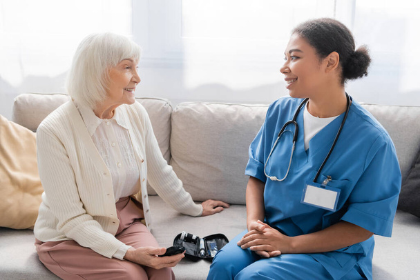 happy senior woman holding glucometer near multiracial nurse in blue uniform  - Photo, Image