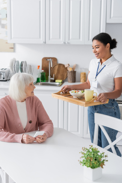 cheerful multiracial social worker holding tray with lunch for senior woman with grey hair  - Photo, Image