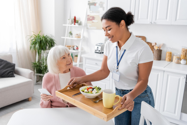 feliz trabajador social multirracial sosteniendo bandeja con almuerzo para mujer mayor con pelo gris  - Foto, imagen