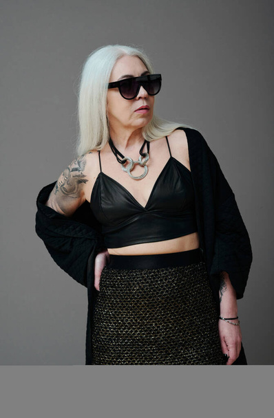 Vertical medium long shot of self-confident mature Caucasian woman wearing fashionable outfit and sunglasses posing on camera, gray background - Photo, Image