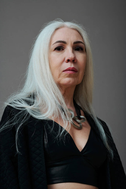 Retrato de estudio de bajo ángulo de atractiva mujer caucásica madura que usa un atuendo de estilo gótico moderno mirando a la cámara, fondo gris - Foto, Imagen