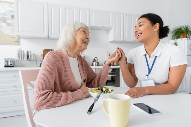 mujer jubilada feliz con el pelo gris cogido de la mano con alegre trabajador social multirracial al lado del almuerzo en la mesa  - Foto, imagen
