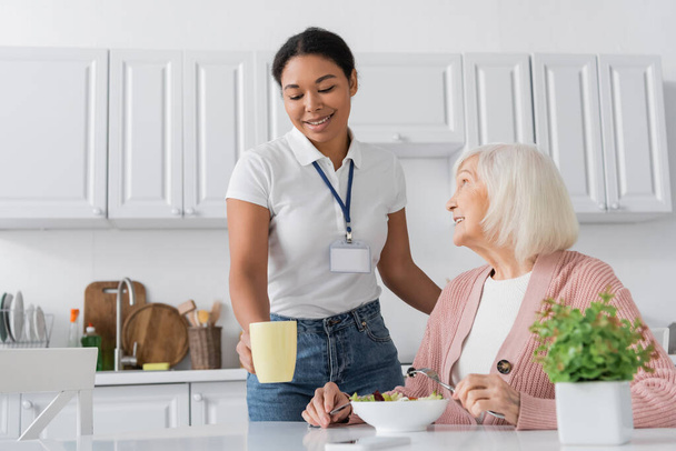 happy multiracial social worker holding cup of tea near retired woman with grey hair having lunch in kitchen  - Foto, Imagem