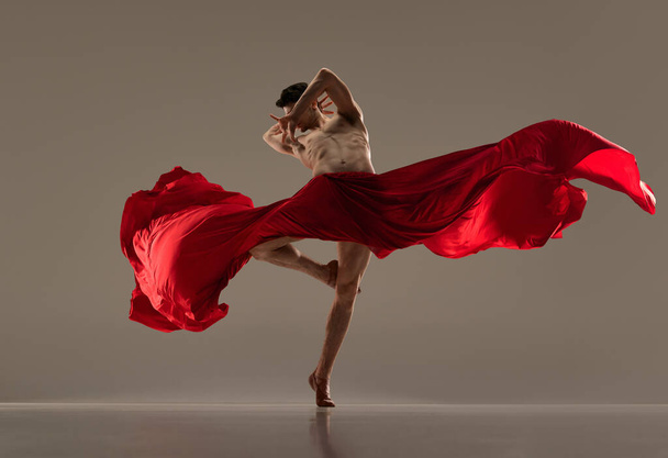 Handsome, passionate man, ballet dancer making impressive performance with red silk fabric against grey studio background. Concept of art, classical dance, inspiration, creativity, choreography - 写真・画像