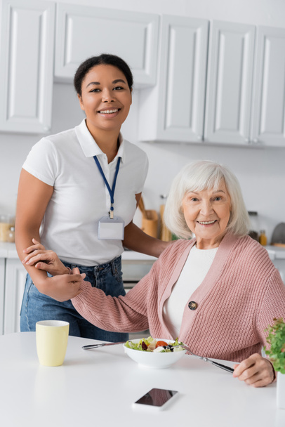 happy multiracial social worker holding hands of thankful retired woman while looking at camera  - Photo, Image