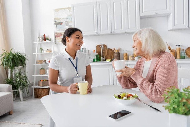 happy multiracial social worker having tea with senior woman in kitchen  - Photo, Image