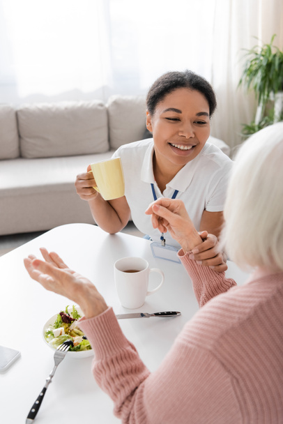 happy multiracial social worker having tea during conversation with senior woman in kitchen  - Valokuva, kuva