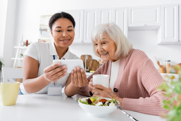 trabajador social multirracial feliz mostrando teléfono inteligente a la mujer jubilada durante el almuerzo en la cocina  - Foto, Imagen