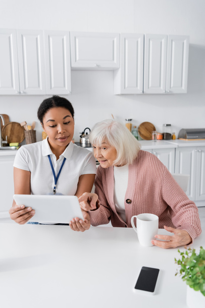 happy multiracial social worker showing digital tablet to senior woman in kitchen  - Foto, afbeelding