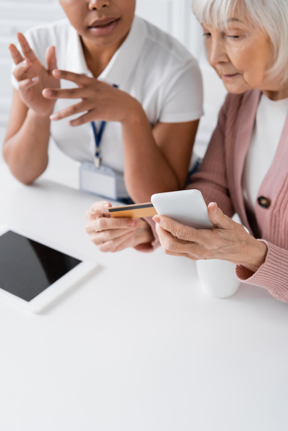 multiracionális szociális munkás elmagyarázza idősebb nő hitelkártyával és okostelefonnal, hogyan kell online vásárlás  - Fotó, kép