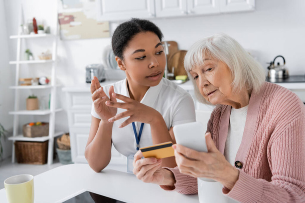 multiracionális szociális munkás elmagyarázza a nyugdíjas nő hitelkártyával és okostelefonnal, hogyan kell online vásárlás  - Fotó, kép