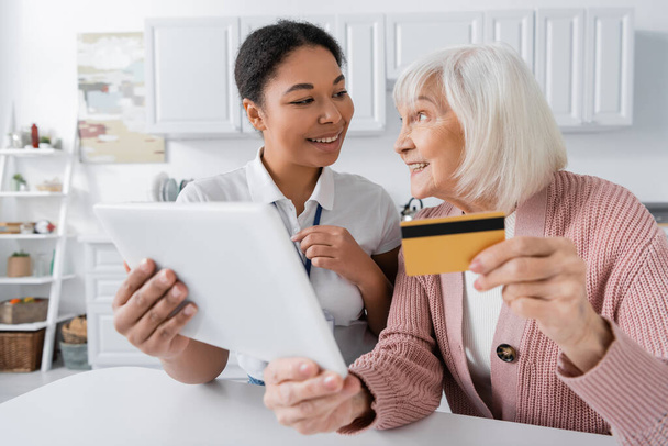 vidám multiracionális szociális munkás digitális táblagép közelében boldog idős nő hitelkártyával  - Fotó, kép