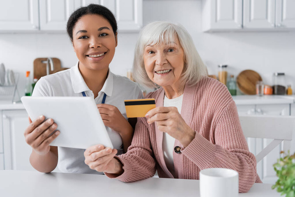 vidám multiracionális szociális munkás digitális táblagépet tart közel pozitív idős nő hitelkártyával  - Fotó, kép