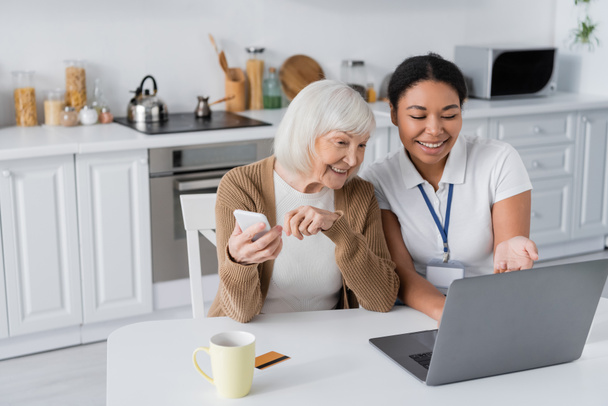 boldog multiracionális szociális munkás használja laptop közelében nyugdíjas nő okostelefon  - Fotó, kép