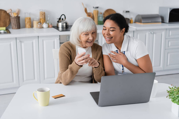 vidám multiracionális szociális munkás néz okostelefon kezében nyugdíjas nő mellett laptop  - Fotó, kép