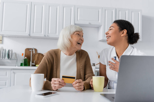 cheerful multiracial social worker holding cup near happy senior woman with credit card and gadgets   - Photo, Image