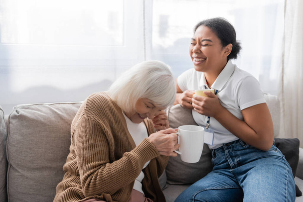 gelukkig multiraciale maatschappelijk werker lachen met senior vrouw terwijl het hebben van thee in de woonkamer  - Foto, afbeelding