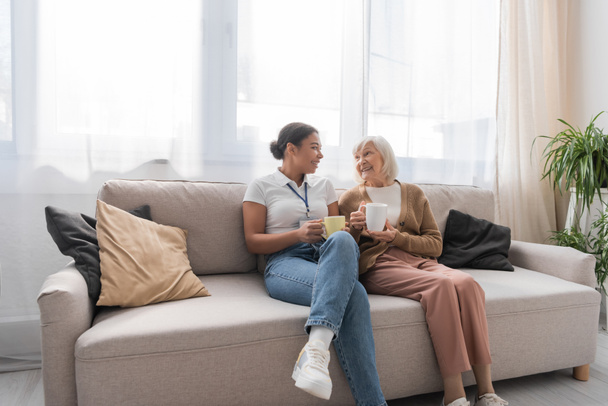 happy multiracial social worker having tea with senior woman in living room  - Photo, Image