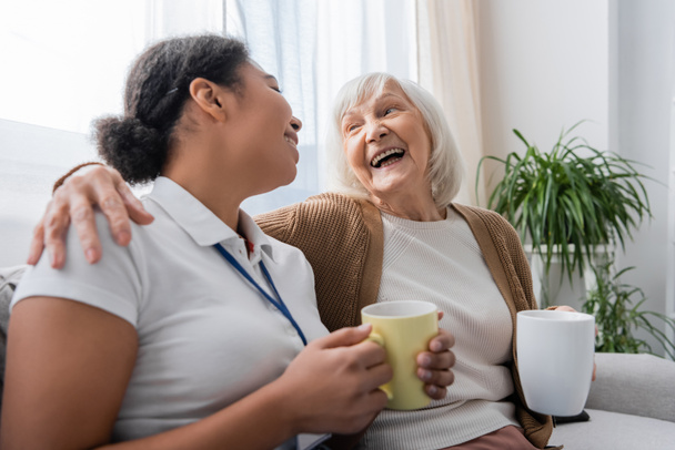 assistente social multirracial feliz tomando chá e rindo com a mulher idosa na sala de estar  - Foto, Imagem