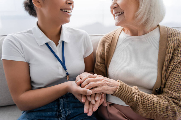 vue recadrée de travailleur social multiracial heureux tenant la main avec une femme âgée dans le salon  - Photo, image
