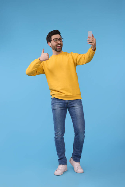 Smiling man taking selfie with smartphone and showing thumbs up on light blue background - Photo, image